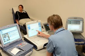 Zorgmedewerker bedient medische apparatuur terwijl patiënt op onderzoeksbank ligt, aangesloten op sensoren.