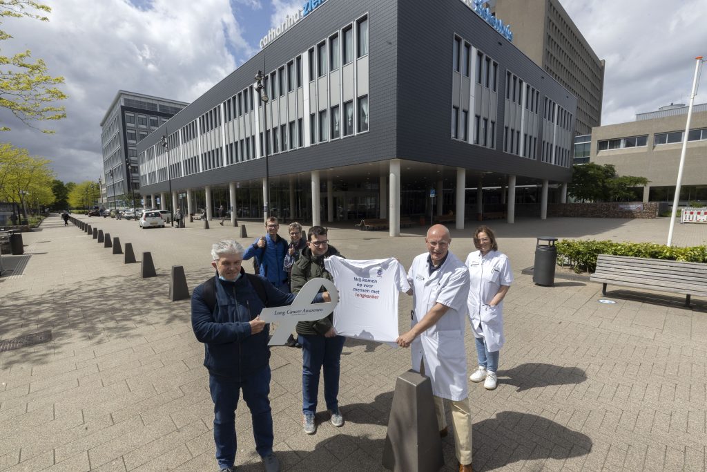 Groep mensen, twee in witte jassen, voor Catharina Ziekenhuis met grote sleutel en "longkanker" T-shirt. De helft bewolkt.