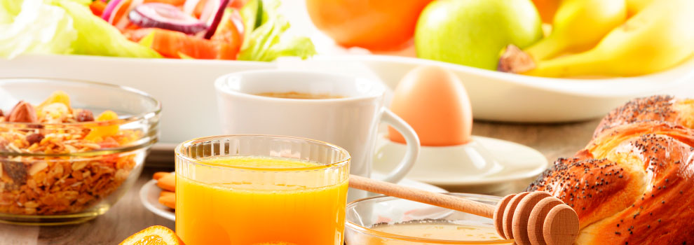 Ontbijt met sinaasappelsap, granola, koffie, gekookt ei, honing, salade, brood en fruit op de achtergrond.