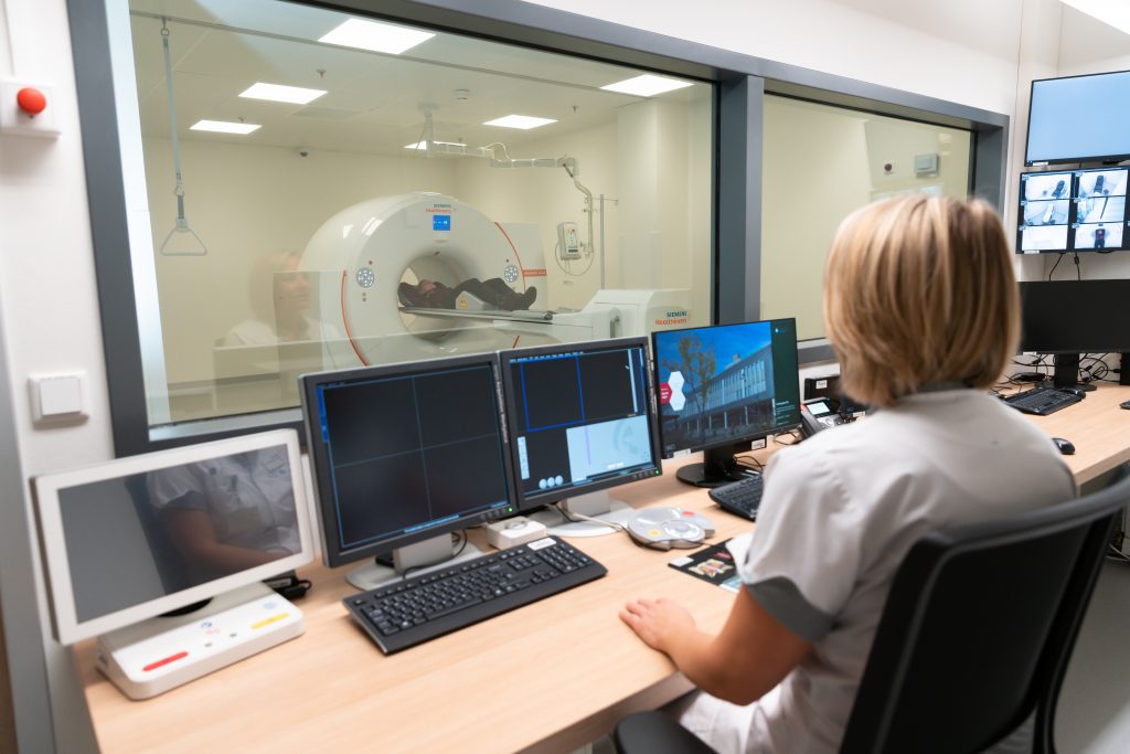 Een medisch technicus in Zuidoost-Brabantse ziekenhuizen bewaakt een CT-scan via een raam en computerschermen.