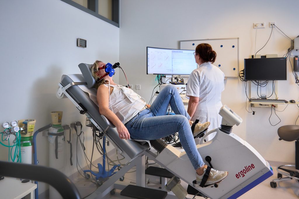 Persoon ligt op een ergoline hometrainer met ademmasker; zorgverlener controleert meetgegevens in onderzoeksruimte.