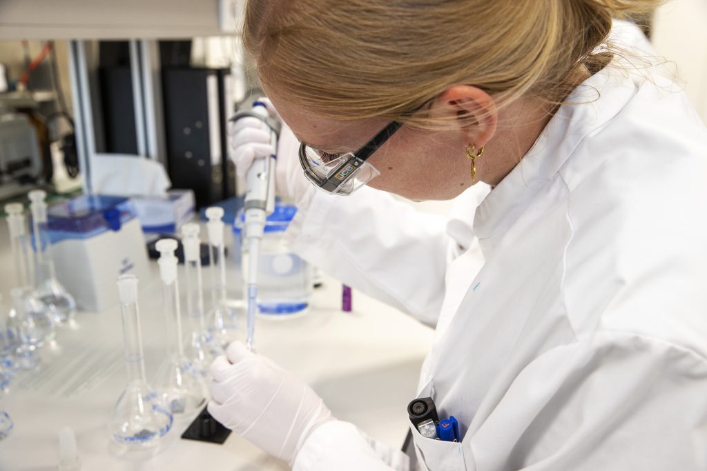 Persoon in witte labjas met handschoenen en bril gebruikt pipet in lab, benadrukt rol van software bij medicatieveiligheid.