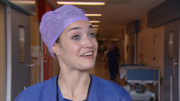 Persoon in scrubs en paarse haardeksel staat glimlachend in ziekenhuisgang, klaar voor opname Borstkanker-voorlichtingsvideo.