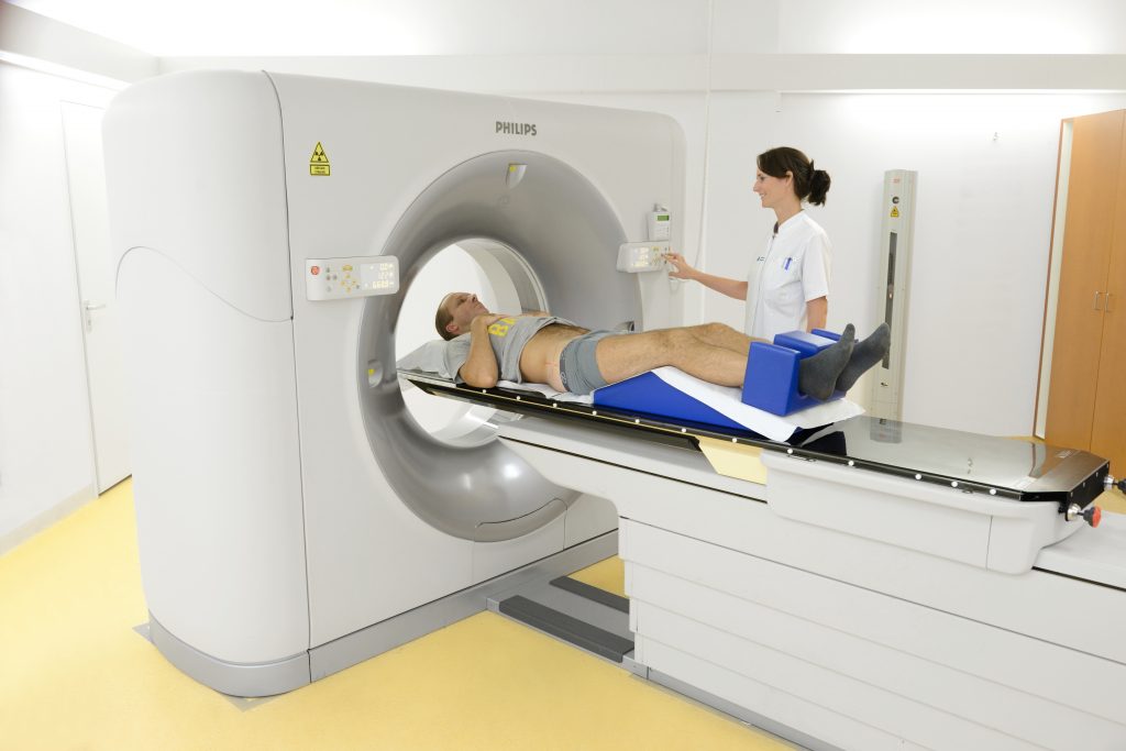 Een persoon ligt op een CT-scanner terwijl een zorgverlener bij het bedieningspaneel in een medische beeldvormingsruimte staat.