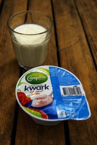 Een glas melk en een dichte Campina kwark met blauwe deksel en aardbeien, op een houten ondergrond.