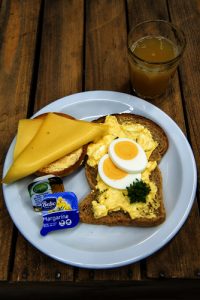 Bord met twee sneetjes brood: één met ei en peterselie, één met kaas. Margarine en sap op de achtergrond.