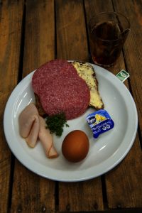 Wit bord met salami op brood, kalkoen, ei, margarine en kruiden. Glas donkere thee ernaast op houten tafel.