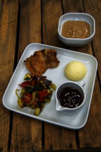 Wit vierkant bord met vlees, aardappelpuree, groenten en worst. Apart bakje met lichtbruin dessert op houten tafel.