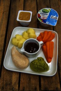 Wit vierkant bord met aardappels, wortels, groentepuree, kipfilet en worst. Yoghurt en appelmoes erbij.