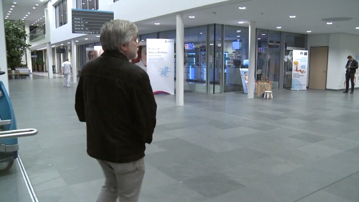 Een man met grijs haar en bril loopt in een ruime lobby van het Catharina Kanker Instituut langs informatieve video's over kanker.
