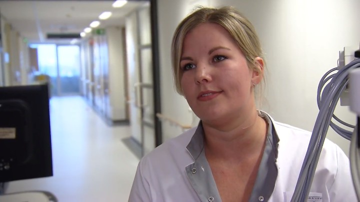 Vrouw in witte jas staat op ziekenhuisgang bij Catharina Kanker Instituut, kijkt voorbij de camera. Apparatuur en monitor zichtbaar.