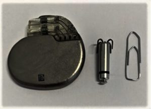 Drie objecten op een witte ondergrond: een pacemaker, een kleine cilinderpacemaker en een paperclip ter groottevergelijking.