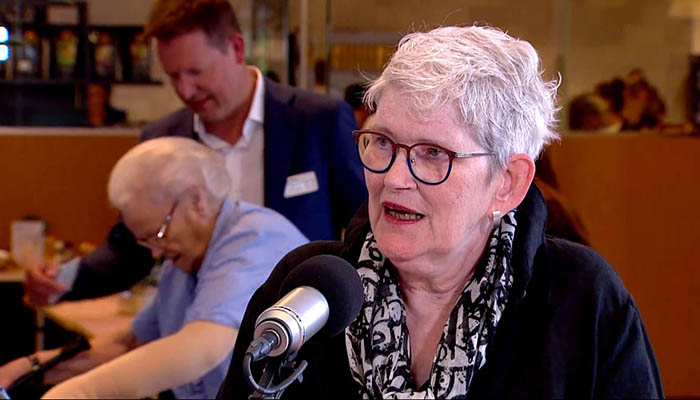 Oudere vrouw met grijs haar en bril spreekt in microfoon. Twee mensen op achtergrond bezig met eigen activiteiten.