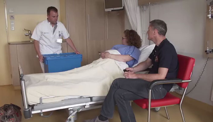 Een anesthesieverpleegkundige staat bij een ziekenhuisbed en spreekt met een patiënt; een man zit in een rode stoel erbij.
