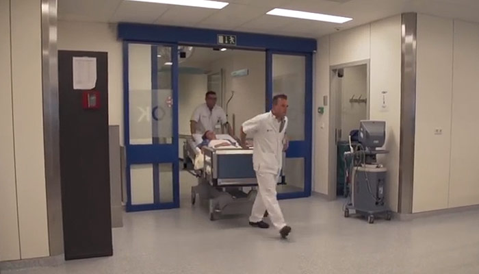 Twee medisch medewerkers in witte uniformen rijden een patiënt op bed door blauwe deuren in een heldere, schone bende.