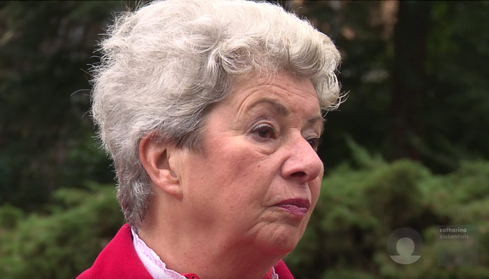 Oudere vrouw met kort, grijs krullend haar buiten in rood jack en witte blouse, kijkt neutraal opzij; vage groene achtergrond.