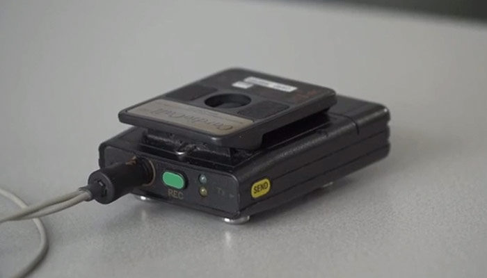 Close-up van een zwarte cassetterecorder met groene REC-knop, gele SEND-knop en kabel op een lichtgrijze ondergrond.