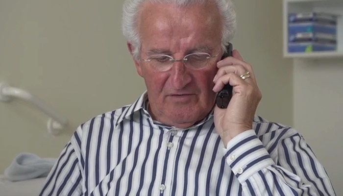 Oudere man met wit haar en bril, in gestreept shirt, riem en kijkt omlaag. Op de achtergrond planken met gestapelde spullen.