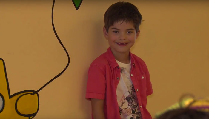 Jonge jongen met kort bruin haar voor gele muur met muurschildering van bier en ballon, glimlacht en draagt roze overhemd.