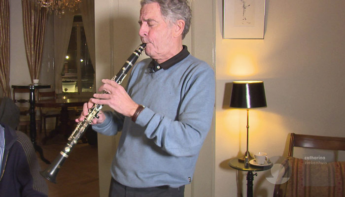Een oudere man in een lichtblauwe trui speelt klarinet in een warm verlichte kamer met lamp, stoel en kopje op een tafeltje.