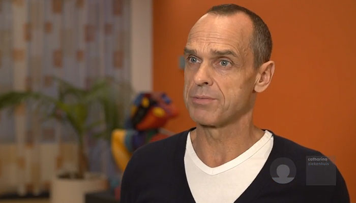 Man met kort haar in donkere trui en wit shirt staat binnen voor oranje muur en raam met gordijn; plant op achtergrond.