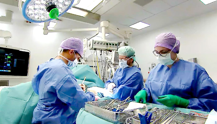 Drie chirurgen in operatiekleding voeren een operatie uit in een verlichte OK met zichtbaar medisch materiaal en apparatuur.