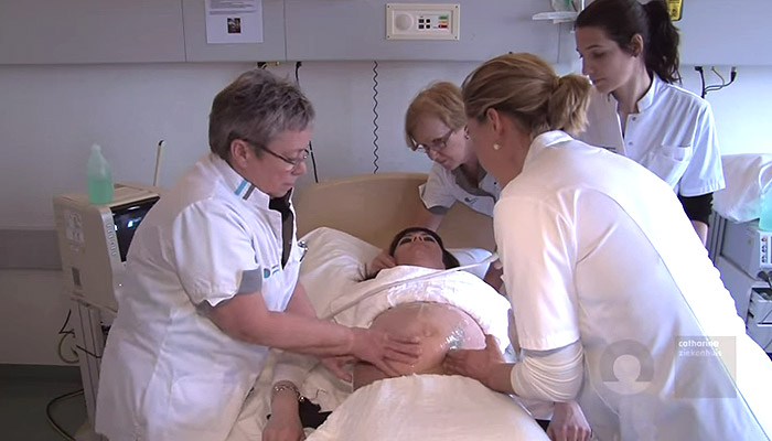 Vier zorgverleners in witte uniformen onderzoeken een zwangere vrouw op een ziekenhuisbed met medische apparatuur in beeld.