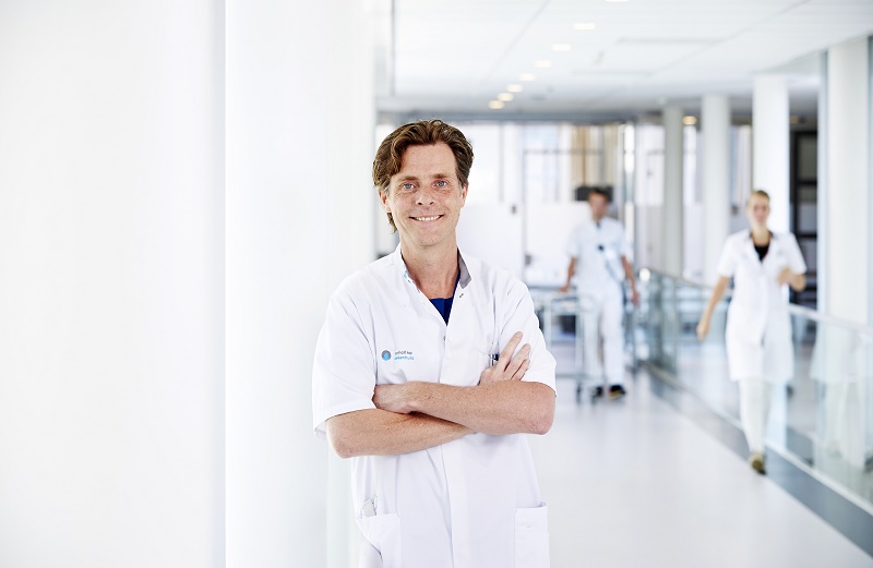 Zorgprofessional in witte jas staat met armen over elkaar in heldere gang; twee collega's lopen op de achtergrond.