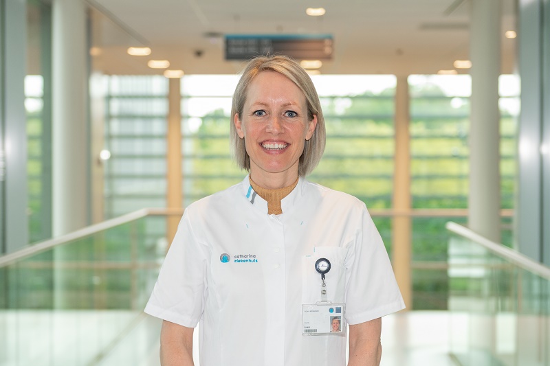 Persoon in witte jas met ID-badge staat in moderne gang met grote ramen; buiten is groen zichtbaar na recente coronamutaties.