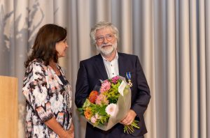 Een vrouw en Harm Rutten met Ridderorde en bloemen staan binnen, kijk elkaar aan. Podium en gordijn op achtergrond.