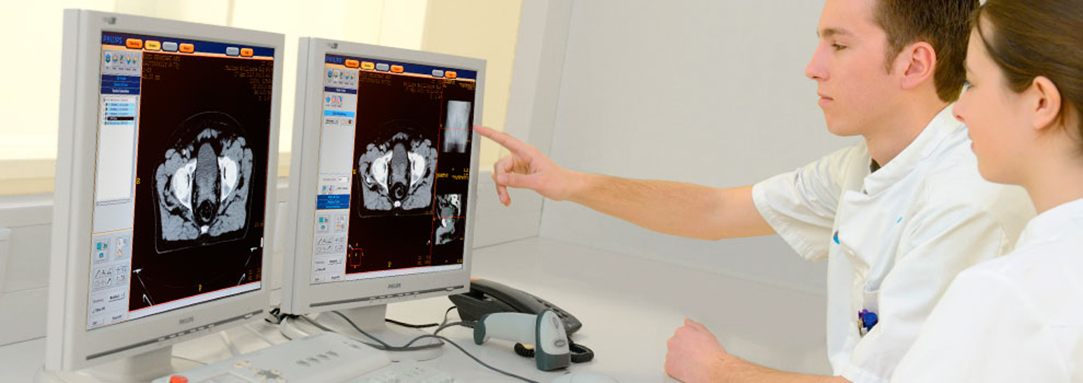 Twee artsen in witte jassen bekijken CT-scans van prostaatkanker op twee schermen; één wijst een gebied aan voor behandeling.