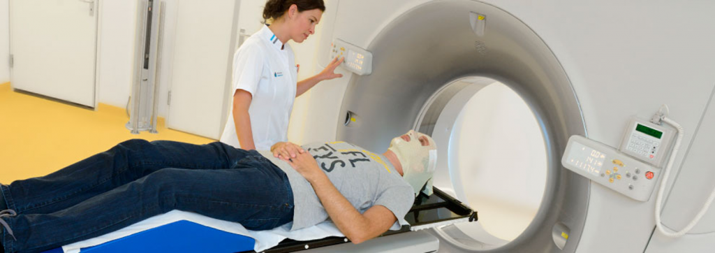 Een patiënt op een tafel gaat een CT-scanner in met hoofdfixatie; een zorgverlener bedient het apparaat en spreekt de patiënt toe.