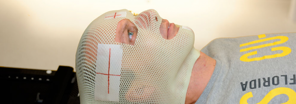 Persoon ligt op rug met wit masker en markeringen voor hoofdbestraling, draagt grijs shirt met gele tekst.