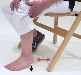 Persoon op houten stoel beweegt blote rechtervoet zijwaarts, vasthoudend aan de knie. Linkervoet draagt een paarse sneaker.