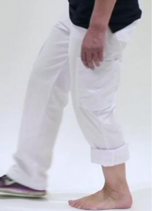 Persoon met witte broek, één pijp opgerold. Linkervoet bloot, rechtervoet met sneaker. Effen lichte achtergrond.
