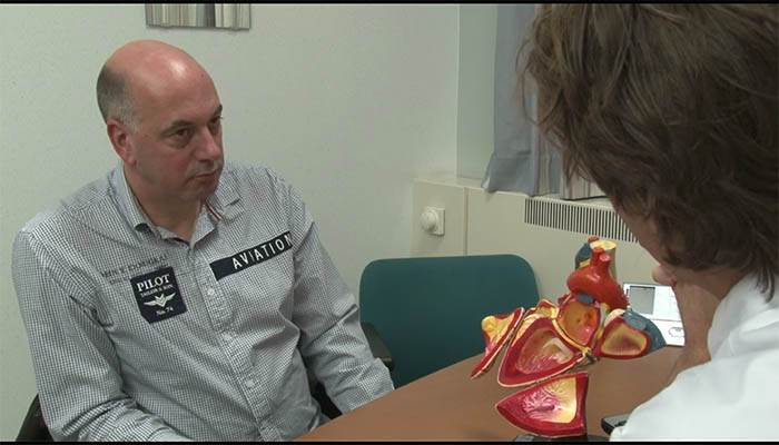 Een man met PILOT- en AVIATION-patches op zijn shirt zit aan een tafel tegenover iemand met een anatomisch hartmodel.