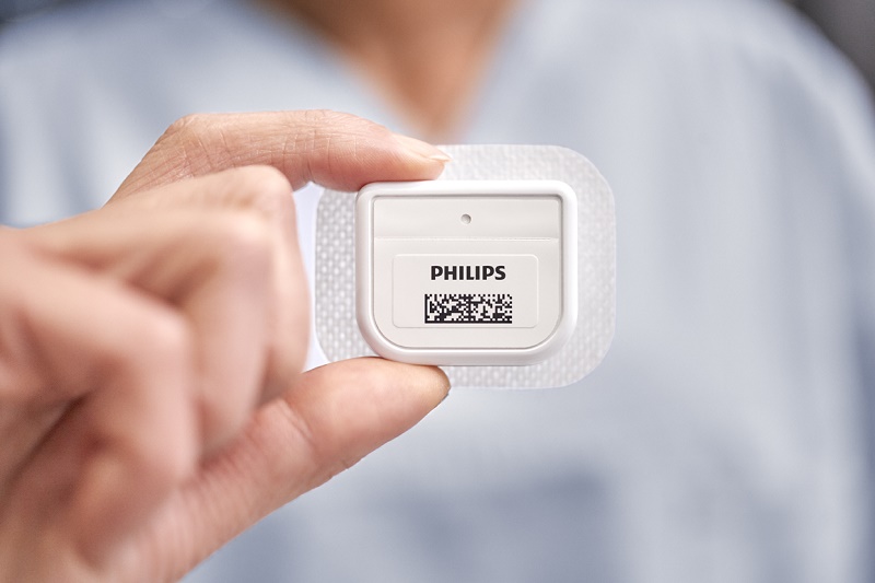 Persoon houdt een rechthoekige Philips slimme sensor met barcode op een vierkante pleister; achtergrond wazig.