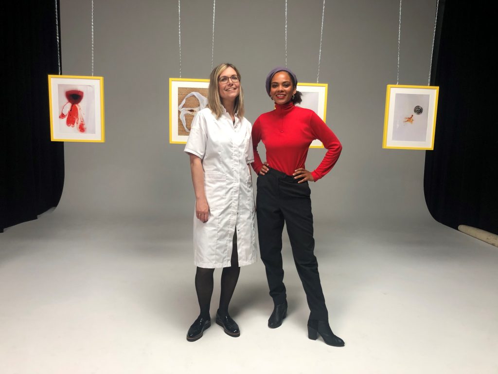Twee vrouwen in een studio met grijze achtergrond; één in een witte jas, één in rood. Vier kunstwerken hangen achter hen.