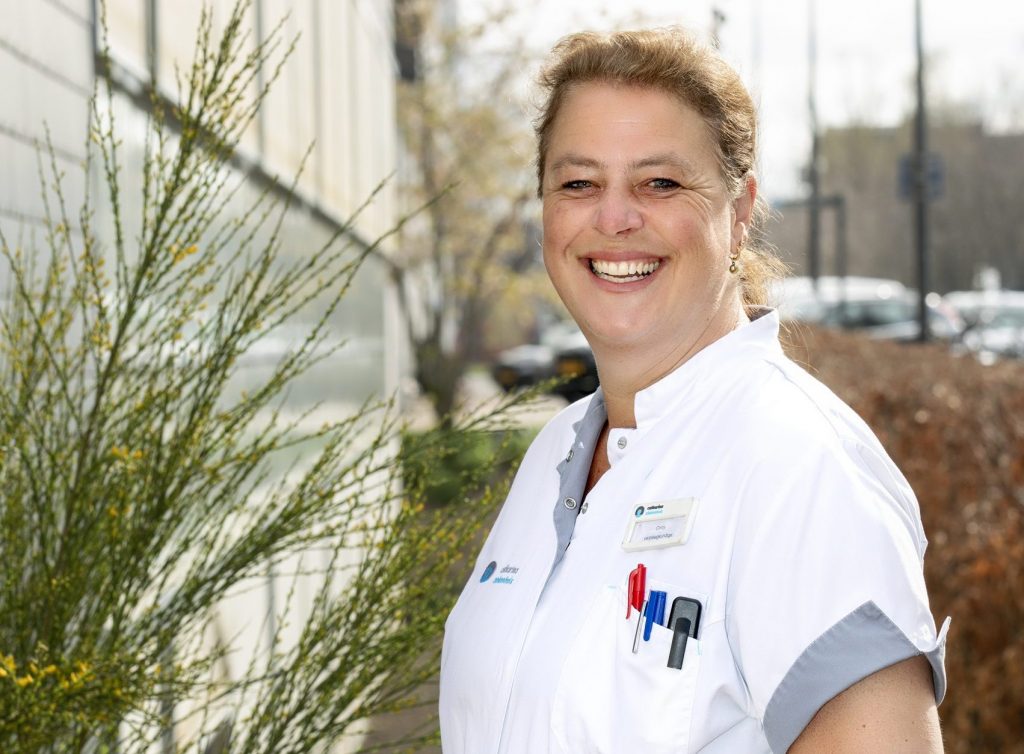 Persoon in witte zorgkleding met naamkaartje "Cindy" staat buiten, lacht. Op achtergrond groen, gebouw en geparkeerde auto's.