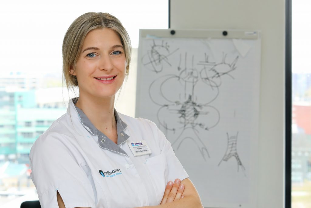Vrouw in witte labjas staat met gekruiste armen en glimlacht, flipover met anatomische tekeningen op de achtergrond, stadsgezicht buiten.