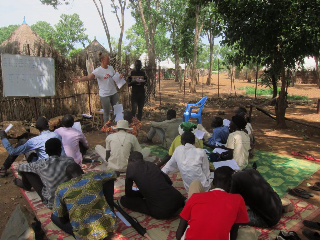 Groep mensen zit op matten buiten in Afrika en luistert naar spreker bij een whiteboard. Op de achtergrond bomen en hutten.