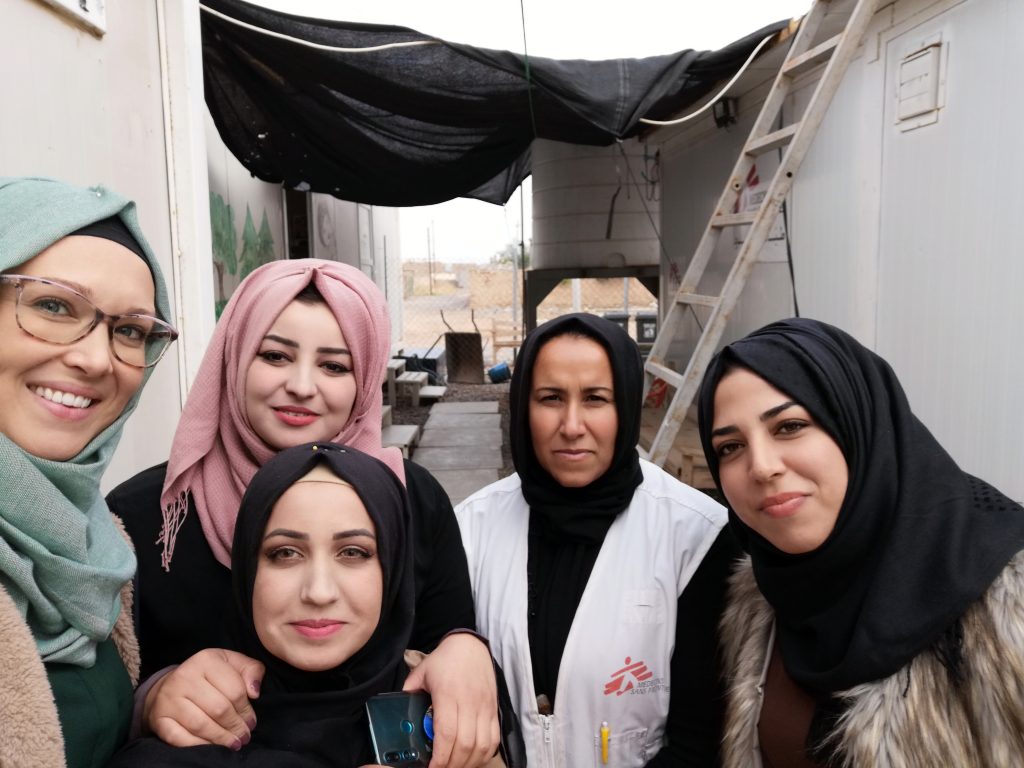 Vijf vrouwen, deels met hoofddoek, eentje in MSF-jas, poseren lachend buiten tussen tijdelijke gebouwen in Irak.