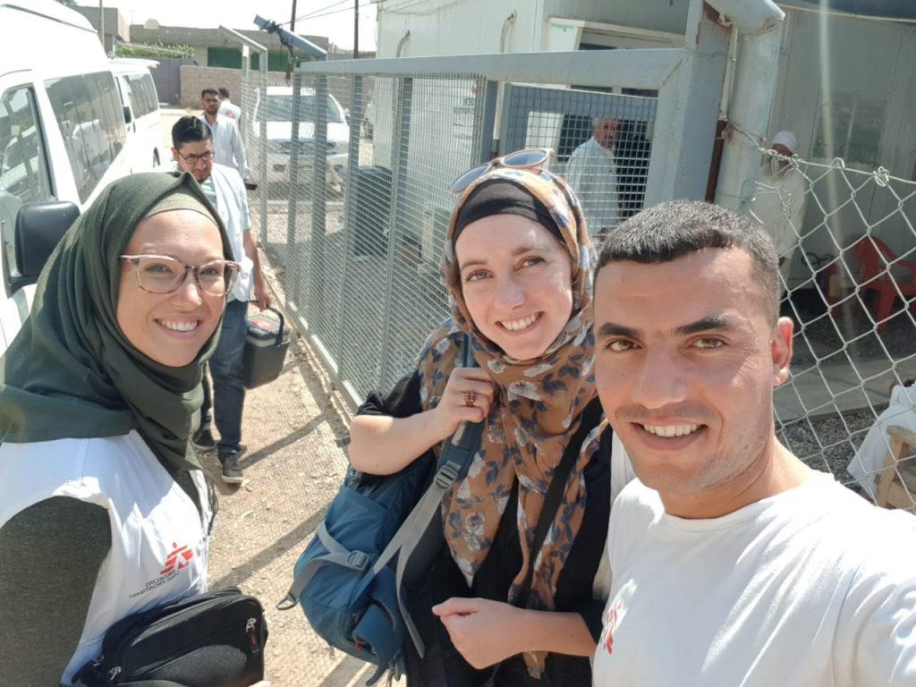 Drie mensen poseren buiten voor een selfie bij een hek in Irak. Twee dragen MSF-shirts, één heeft een blauwe tas.