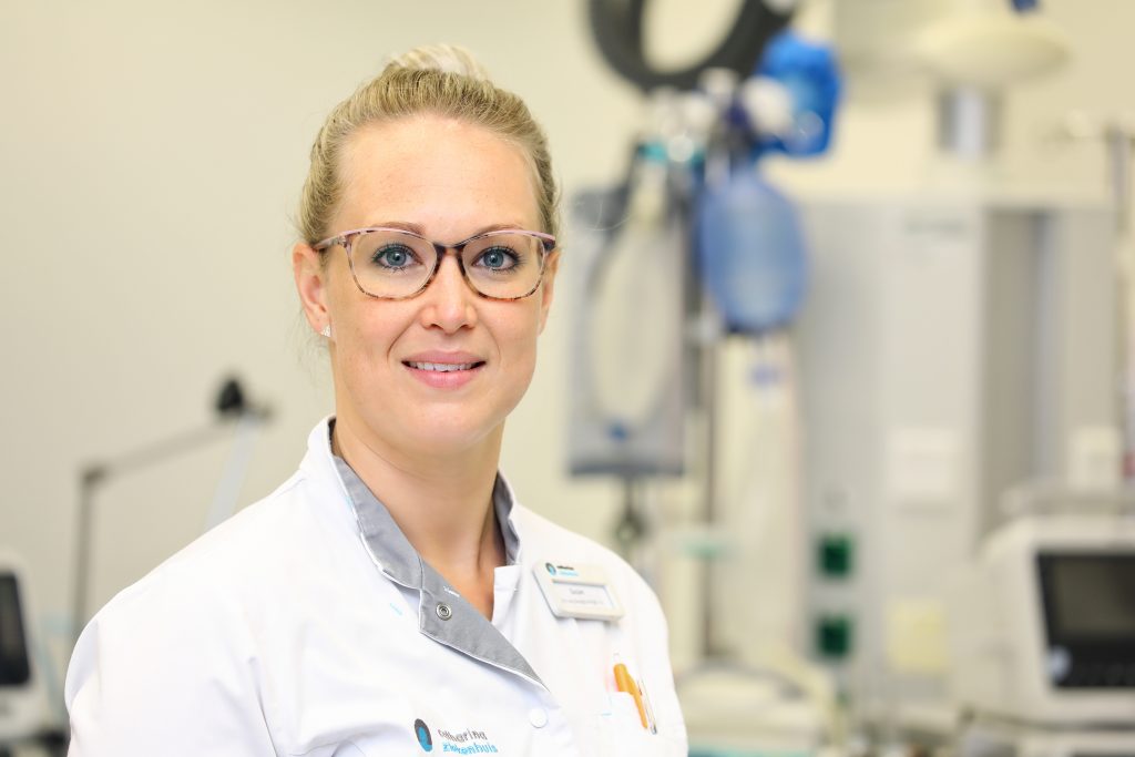 Vrouw met bril en witte labjas in medische omgeving, met apparatuur op de achtergrond. Toegewijd aan zorg van Afrika tot Irak.