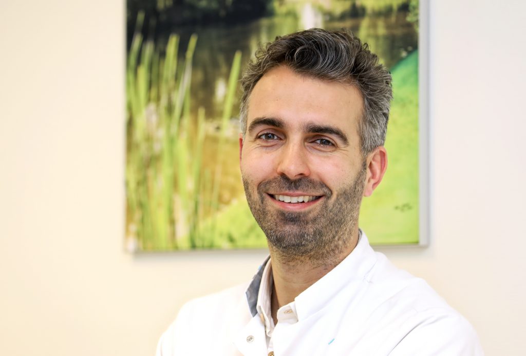 Een man met kort donker haar en een baard glimlacht in een witte jas. Vage planten en water op de achtergrond.