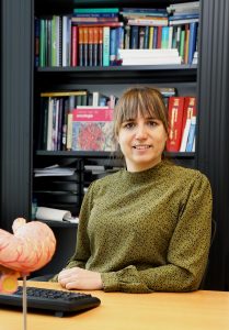 Vrouw met bruin haar en pony zit aan bureau met toetsenbord, maagmodel en boekenplanken. Ze draagt een groene blouse en glimlacht.