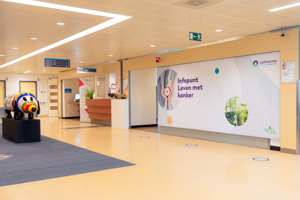 Ziekenhuisgang met balie, wegwijzers, gekleurd koeienbeeld en bord 'Infopunt Leven met kanker', focus op veiligheid en kwaliteit.