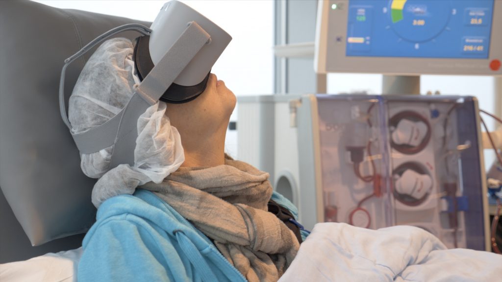 Persoon in medische stoel met haarnet, sjaal en VR-bril; medische apparatuur en monitor op achtergrond in ziekenhuis.
