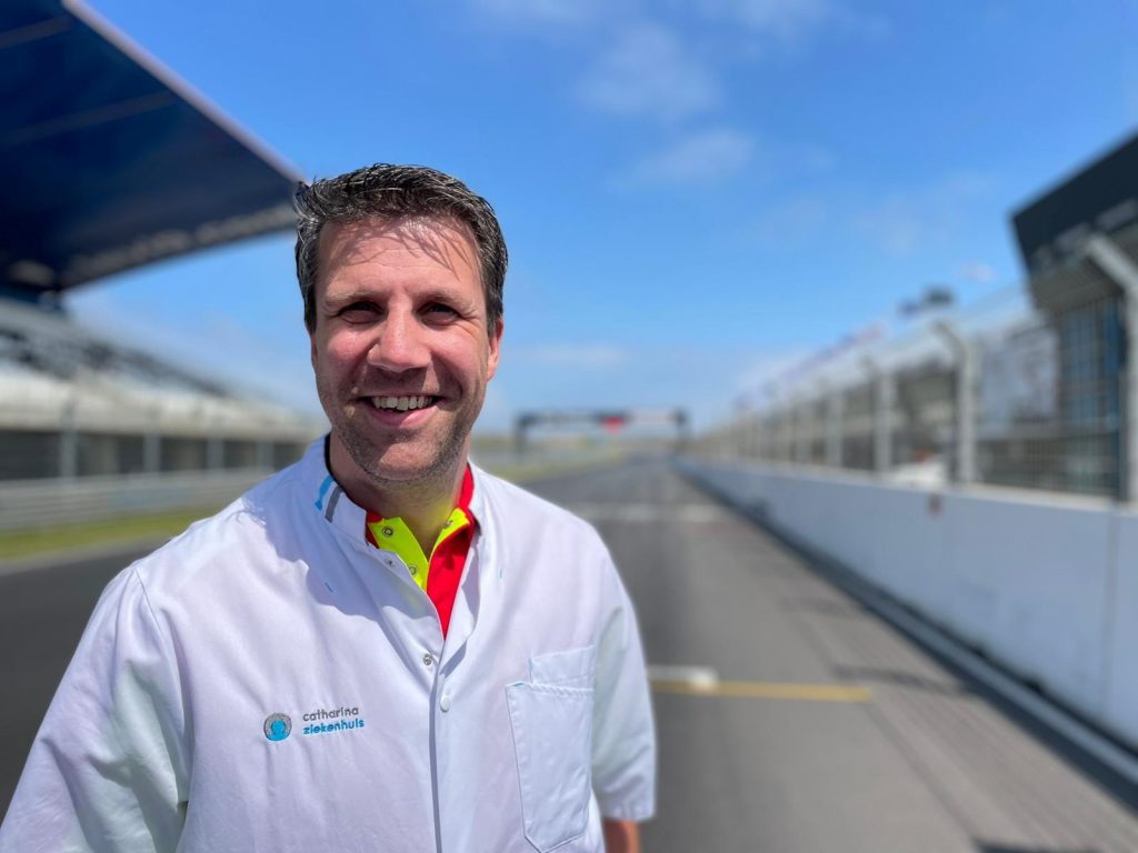 Een lachende anesthesioloog in witte jas staat op het circuit van Zandvoort met lege tribunes en een blauwe lucht.