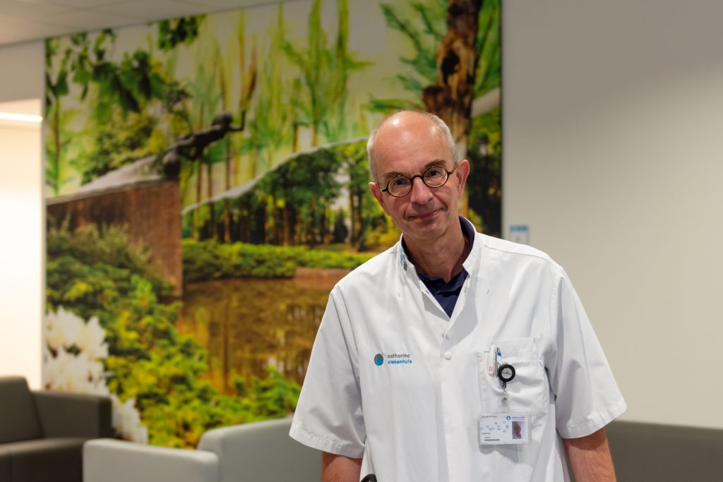 Man met bril en witte labjas staat in moderne ruimte met een natuurmuurschildering, badge zichtbaar aan zijn jas.